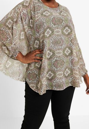 Femme portant un chemisier ample et transparent avec un motif orné vert, marron et rose, associé à un pantalon noir ajusté.