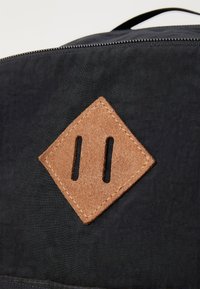 Sac à dos en tissu noir avec un patch de cuir marron en forme de diamant, comportant deux fentes et des coutures contrastées. Texture lisse avec une fermeture éclair.