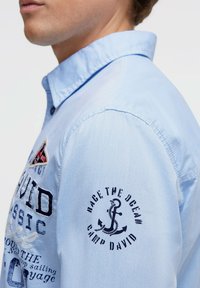 Hellblaues Hemd mit Knöpfen, bestickt mit Details, einer aufgesetzten Tasche und einem runden Logo auf dem Ärmel mit Ankergrafiken und Text.