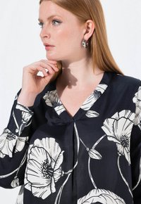 Mustan värinen paita, jossa on suuria valkoisia kukkakuoseja. V-kaula-aukko ja pitkät hihat. Kangas näyttää sileältä ja silkkiseltä, ja se on rento istuvuus.