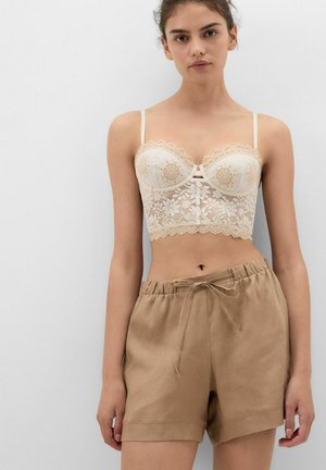 Jonge vrouw draagt een beige kanten crop top met bloemenpatronen en beige shorts met trekkoord, staand tegen een effen witte achtergrond.