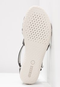 Sandaalit, joissa on pyöreä pitopinta, ilmanvaihtorei'ät ja kohokuvioitu "GEOX"-logo, kallistettuna valkoista kulmaa vasten.