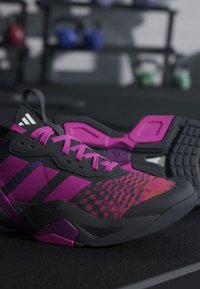 Μαύρα και μωβ αθλητικά παπούτσια Adidas με ροζ κορδόνια σε πάτωμα γυμναστηρίου, ο εξοπλισμός γυμναστηρίου είναι θολός στο φόντο.