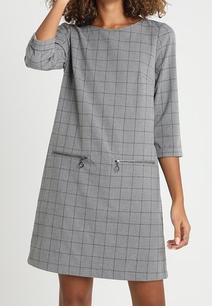 Vrouw draagt een grijze jurk tot op de knie met een ruitpatroon, driekwart mouwen en twee voorzakken met rits.