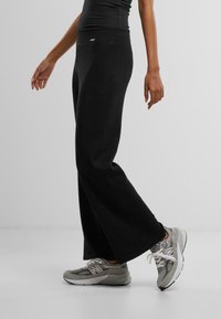 Schwarze weite Hosen mit hohem Bund, aus weichem Stoff gefertigt. Kombiniert mit grauen Sportschuhen, die über Netz- und Gummidetails verfügen.