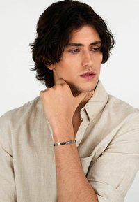 Bracciale d'argento con accenti neri orizzontali, indossato al polso. Chi lo indossa ha capelli scuri e disordinati ed è vestito con una camicia leggera beige.