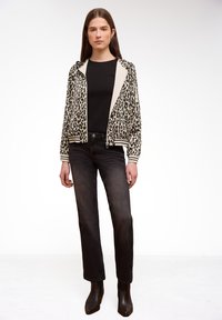 Junge Frau trägt eine Leopardenmuster-Reißverschlussjacke über einem schwarzen Shirt, verblasste schwarze Jeans und schwarze Stiefeletten und steht vor einem weißen Hintergrund.