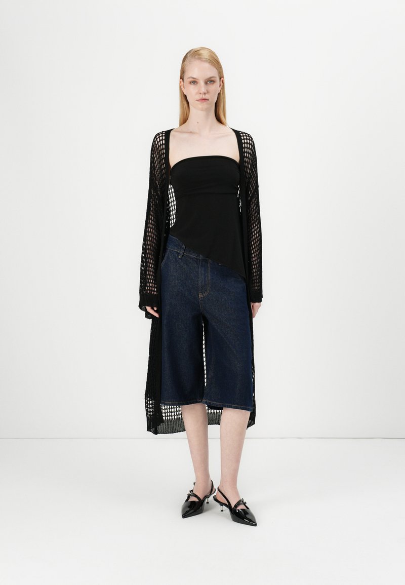 Cárdigan longo de malha preta sobre um top justo preto, combinado com culottes largas de denim azul e sapatos de salto agudo pretos. Design simples e moderno.