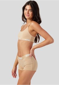 Beige sømløs sports-BH og shorts-sett med hvit logo på midjebåndene, som har et minimalistisk design og myk tekstur.