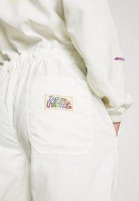 Persoon in witte broek en jasje, met kleurrijke "Love Unites" patch op de achterzak en hand in de zijzak.