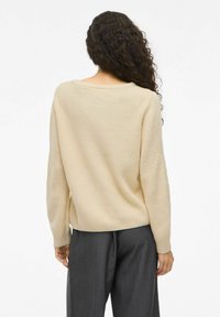 Pull en tricot beige avec une coupe décontractée, doté d'un large col et d'une texture côtelée, associé à un pantalon gris.