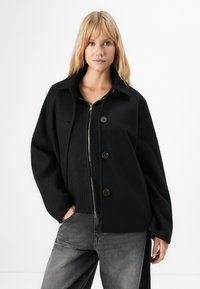 Blond kvinne iført en svart oversized knapp-up frakk over en glidelås svart genser og grå jeans, stående med en hånd i lomma.