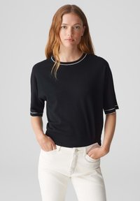 Schwarzer Kurzarmpullover mit rundem Ausschnitt und weißen Akzenten. Kombiniert mit weißen Jeans, die eine lockere Passform und glatte Textur aufweisen.