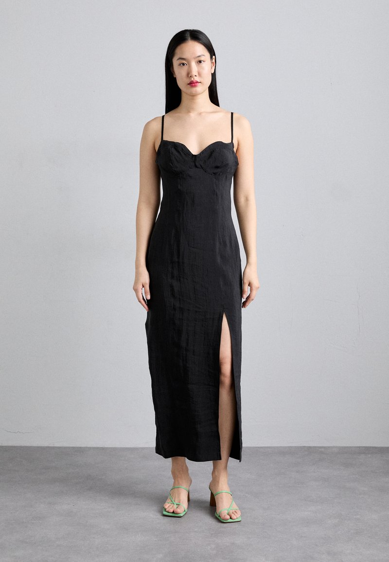 Robe midi en lin noir avec un corsage ajusté, des bretelles fines et une fente sur le côté. Le modèle porte des sandales vertes à lanières. Texture lisse et lignes épurées.