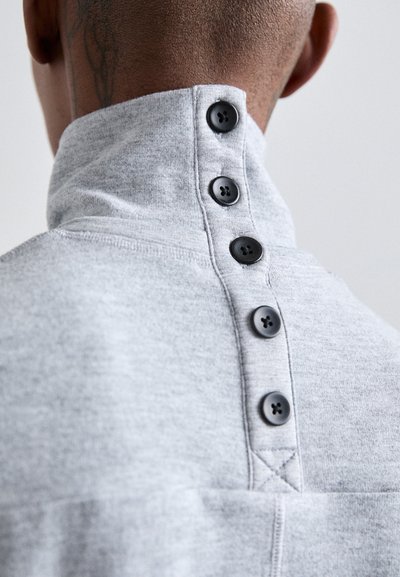 Grijze sweatshirt met een hoge kraag en vijf zwarte knopen aan de achterkant. Zachte textuur, zichtbare stikseldetails en een getailleerde pasvorm.