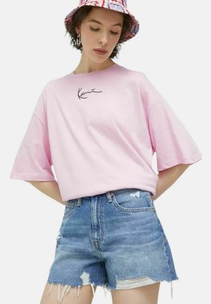 Camiseta de manga corta rosa con un estampado de firma en negro, combinada con pantalones cortos de denim desgastados. La tela parece suave y casual.