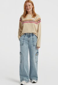 Beige long-sleeve shirt with maroon, orange, and pink stripes; blue wide-leg denim cargo pants; white sneakers. Logo patch on shirt.