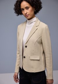 Blazer beige texturé à la coupe ajustée, avec des boutons noirs, un col cranté et deux poches avant. Porté sur un haut blanc.