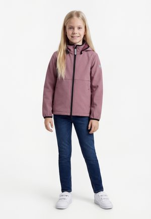 Mlada deklica z dolgimi blond lasmi, oblečena v vijoličasto kapucar jakno, temno modre kavbojke in bele superge z ježki, stoji in se smeji.