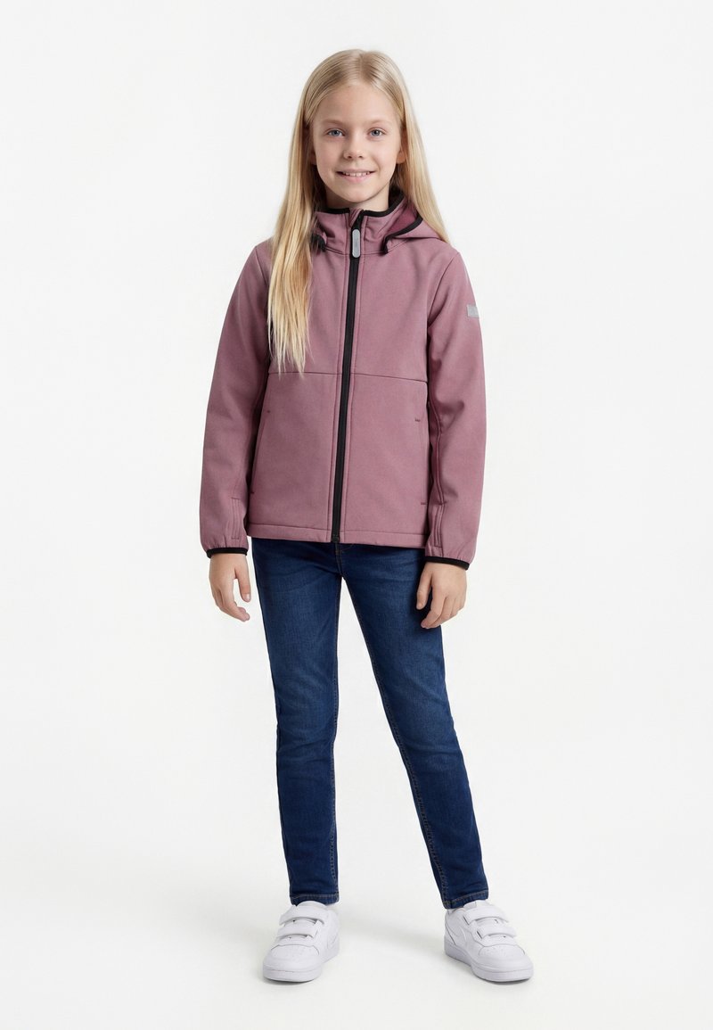 Νεαρή κοπέλα με μακριά ξανθά μαλλιά, φορώντας ένα μωβ μπουφάν με κουκούλα, σκούρο μπλε τζιν και λευκά παπούτσια με βέλκρο, στέκεται και χαμογελάει.