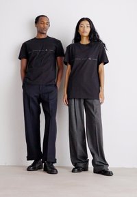 Dos modelos están de pie uno al lado del otro vistiendo camisetas de algodón negras con un detalle de logo. El modelo masculino las combina con pantalones oscuros, la modelo femenina con pantalones grises.