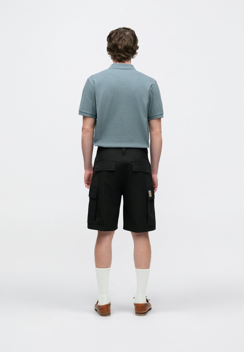 Polo bleu clair associé à un short cargo noir. Le short comporte plusieurs poches et une étiquette de marque visible. Chaussettes blanches et chaussures marron.