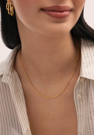 Primer plano del cuello con un delicado collar de cadena dorada y pendientes de aro dorados, vistiendo una camisa beige con cuello y finas rayas verticales.