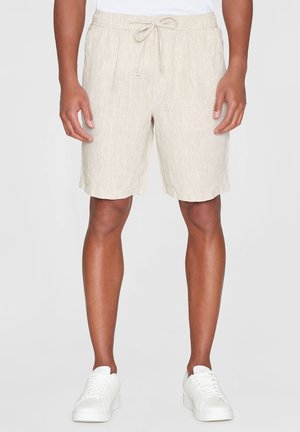 Beige Leinenshorts mit elastischem Bund und Kordelzug. Mit seitlichen Taschen und einer lockeren Passform, kombiniert mit weißen Sneakers.