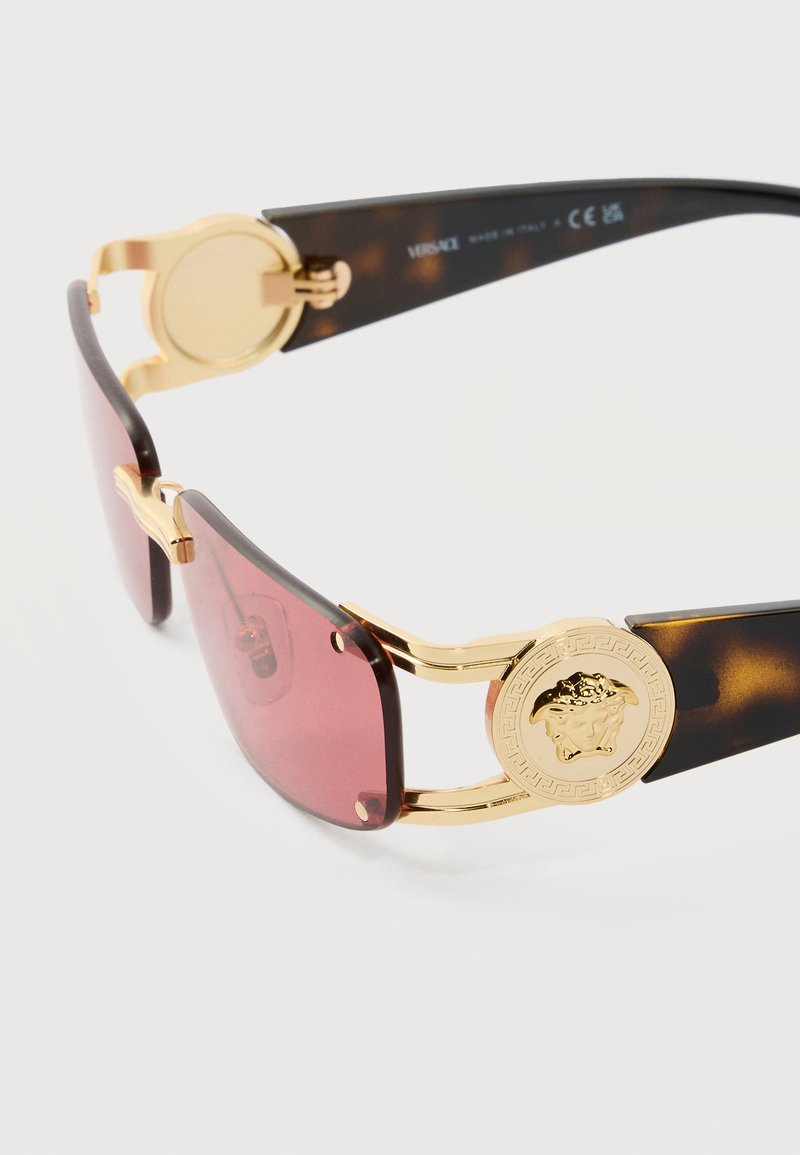 Lunettes de soleil avec des verres rectangulaires marron, des branches en écaille de tortue, et des médaillons Medusa dorés sur les charnières.