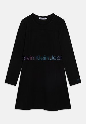 Zwarte jurk met lange mouwen en ronde hals, met een regenboogkleurige "Calvin Klein Jeans" logoband bij de taille en een kleine "ck" logo op de linkermouwmanchet.