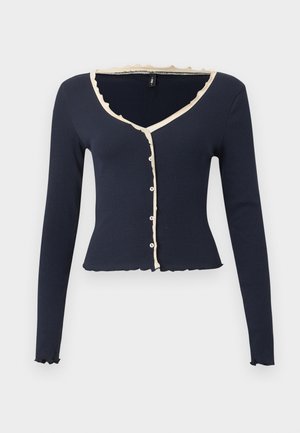 Μακρυμάνικο cropped navy ζακέτα με μπεζ βολάν, V-λαιμόκοψη και μπροστινά κουμπιά σε απλό ανοιχτό φόντο.