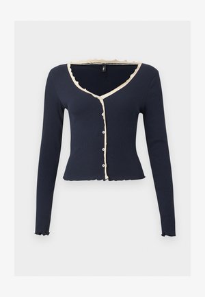 Μακρυμάνικο cropped navy ζακέτα με μπεζ βολάν, V-λαιμόκοψη και μπροστινά κουμπιά σε απλό ανοιχτό φόντο.