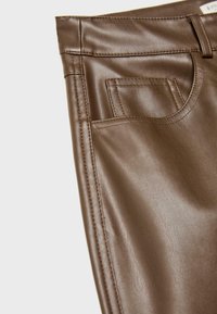 Gros plan sur un pantalon en cuir synthétique marron montrant la ceinture, les passants de ceinture et les détails des poches avant cousues.