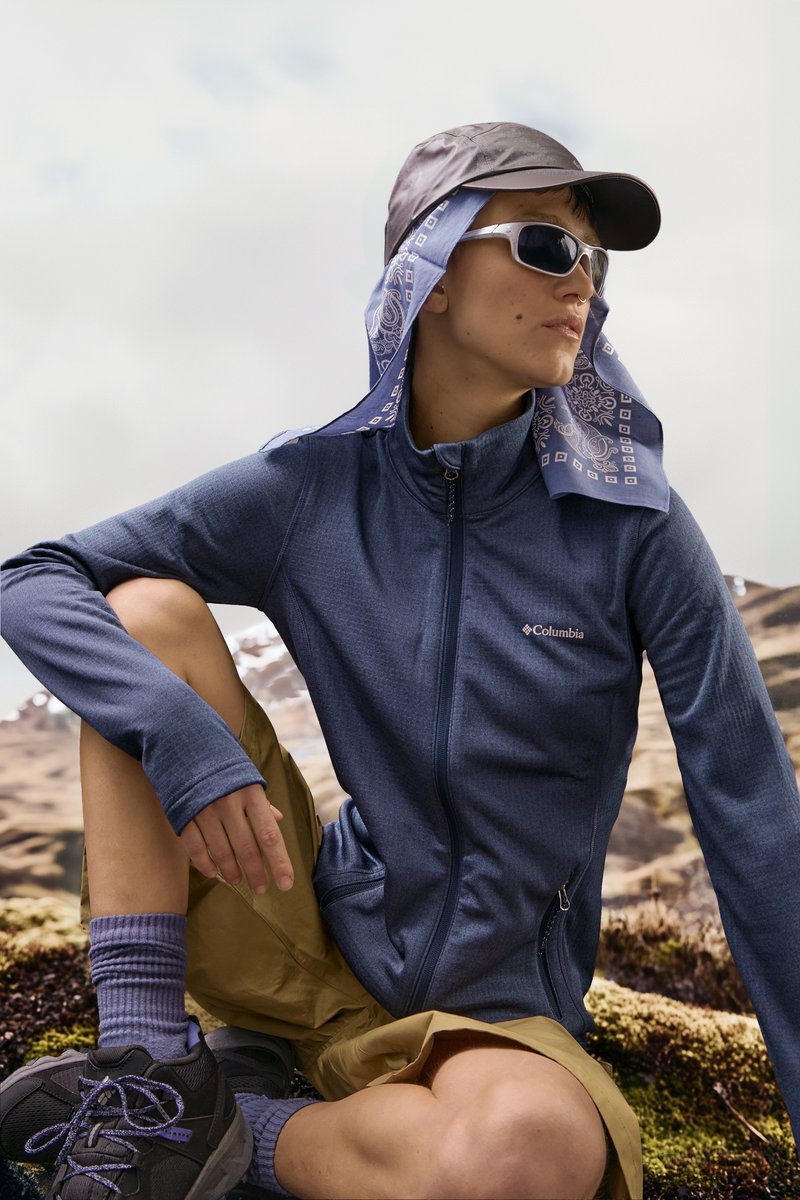 Persoon in een Columbia-jack, met een pet met blauwe bandana, zonnebril, beige shorts en wandelschoenen, die buiten rust neemt op een bergachtig pad.