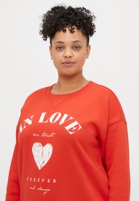 Raudonas džemperis su apvalia iškirpte, ilgomis rankovėmis ir baltu spaudiniu su širdies piešiniu. Minkšta medžiaga, laisvas pasijungimas.