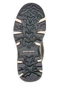 Semelle de chaussure en caoutchouc noir avec motifs de grip texturés et accents beige. Présente des crampons profonds pour la traction et un logo en relief au centre.