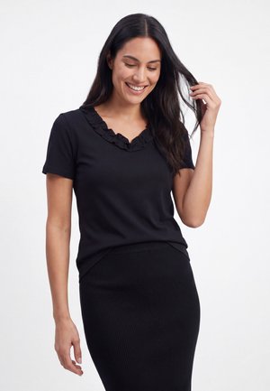 Femme portant un haut noir à manches courtes avec encolure à volants et une jupe noire côtelée, souriant en touchant ses cheveux.
