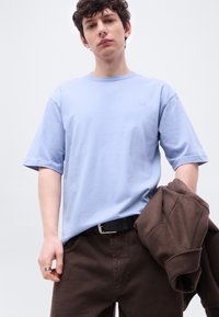 Νέος άνδρας που φοράει ανοιχτό μπλε μπλουζάκι και σκούρο καφέ παντελόνι, κρατώντας ένα διπλωμένο σκούρο καφέ σακάκι στο αριστερό του χέρι μπροστά σε απλό φόντο.