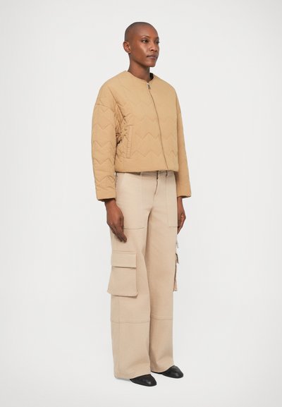 Giacca bomber trapuntata beige con colletto rotondo e chiusura con zip, abbinata a pantaloni cargo chiari con tasche laterali e vestibilità rilassata.