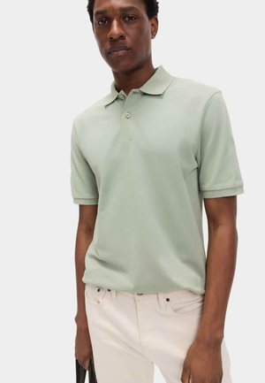 Polo de color verde pálido hecho de tela suave, con cuello clásico, tapeta de dos botones y mangas cortas, combinado con pantalones de color claro.