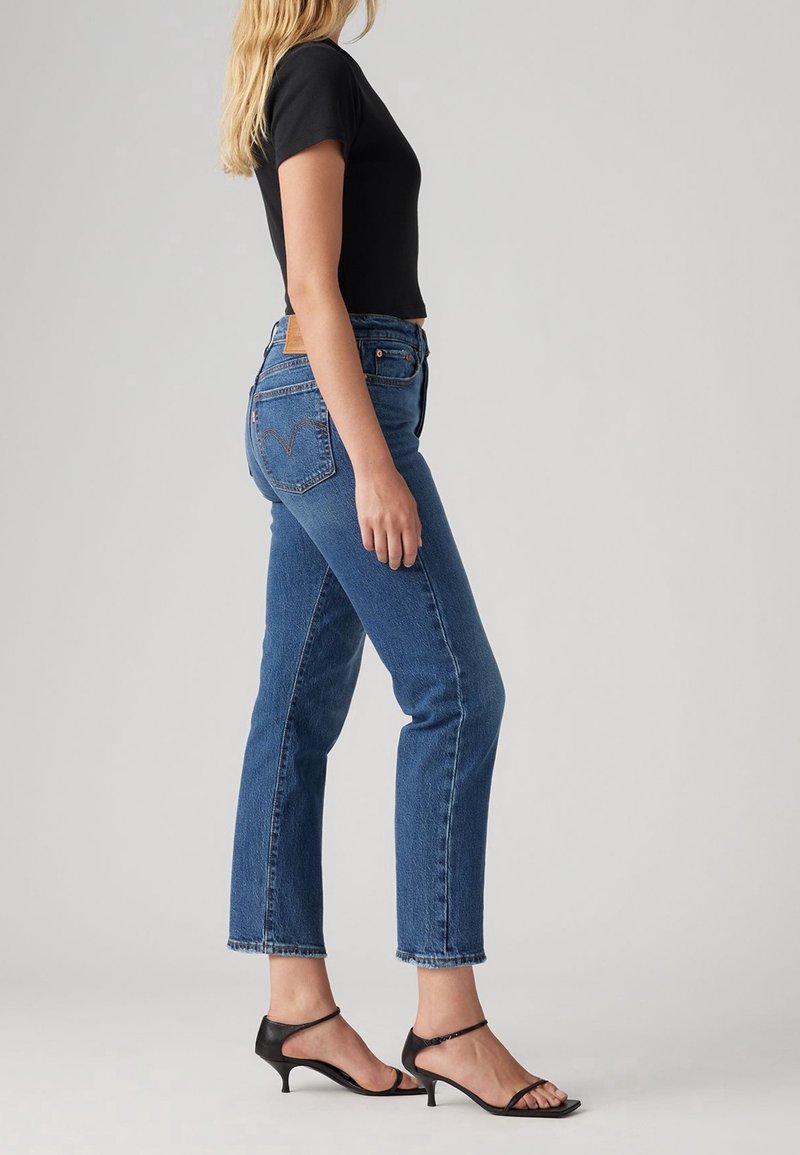 Femme de profil portant un jean droit taille haute bleu, un top noir à manches courtes et des sandales à talons noires sur fond uni.