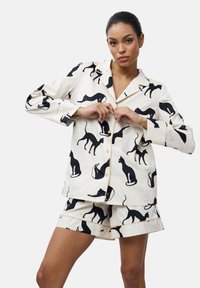 Pigiama di cotone bianco con motivo di gatti neri, composto da una camicia con bottoni e pantaloni corti, rifinito con piping chiaro e una vestibilità comoda.