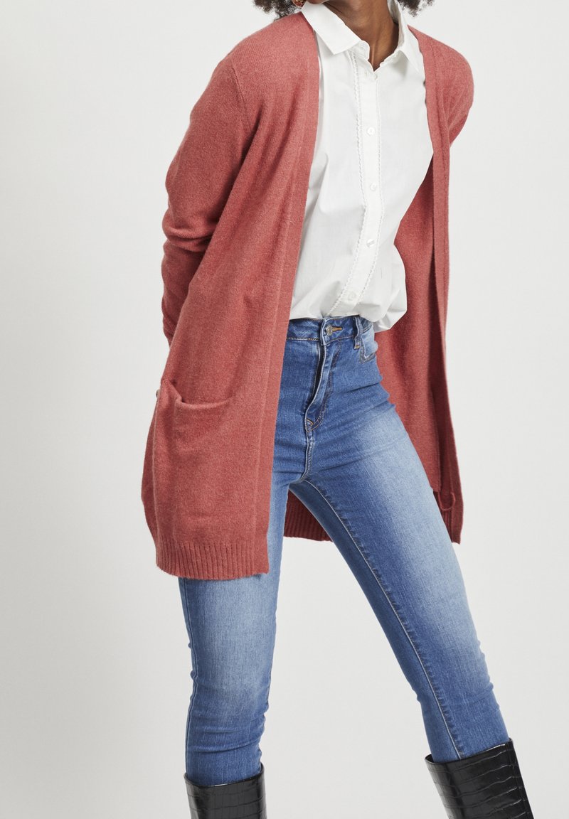 Personne portant un cardigan couleur rouille sur un chemisier blanc, un jean bleu et des bottes noires montantes jusqu’aux genoux sur un fond uni.