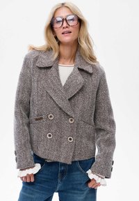 Kobieta w oversize'owej brązowej marynarce w jodełkę, białych rękawach z falbanami, dużych kwadratowych okularach przeciwsłonecznych, w niebieskich dżinsach, stojąca z rękami w kieszeniach.