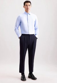 Lichtblauw overhemd met lange mouwen, dichtgeknoopt, gecombineerd met donkerblauwe broek en zwarte nette schoenen, allemaal op een neutrale achtergrond.