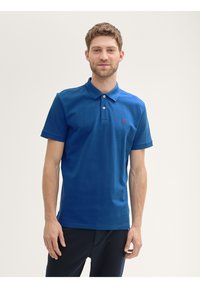 Koszulka polo z krótkim rękawem w niebieskim kolorze, wykonana z bawełny, z zapięciem na dwa guziki i małym czerwonym logo na klatce piersiowej. Gładka tekstura.