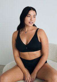 Ensemble de lingerie noire comprenant un bralette à décolleté en V et une culotte taille haute avec des accents transparents. Tissu lisse, sans matériel visible.