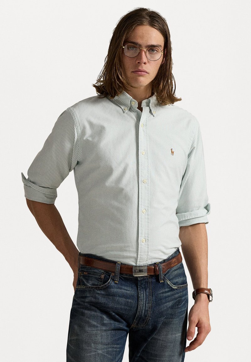 Chemise à rayures vert clair et blanche avec un petit logo sur la poitrine, associée à un jean bleu foncé et une ceinture en cuir marron.