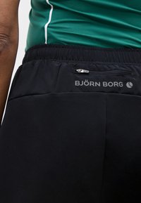 Κοντινό πλάνο ατόμου που φοράει μαύρα σορτς Björn Borg με φερμουάρ στην πίσω τσέπη και πράσινο αθλητικό τοπ με λευκή επένδυση.