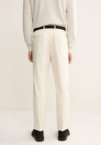 Pantalones beiges con un corte ajustado, que presentan bolsillos traseros y pliegues verticales, combinados con un cinturón negro y zapatos oscuros.